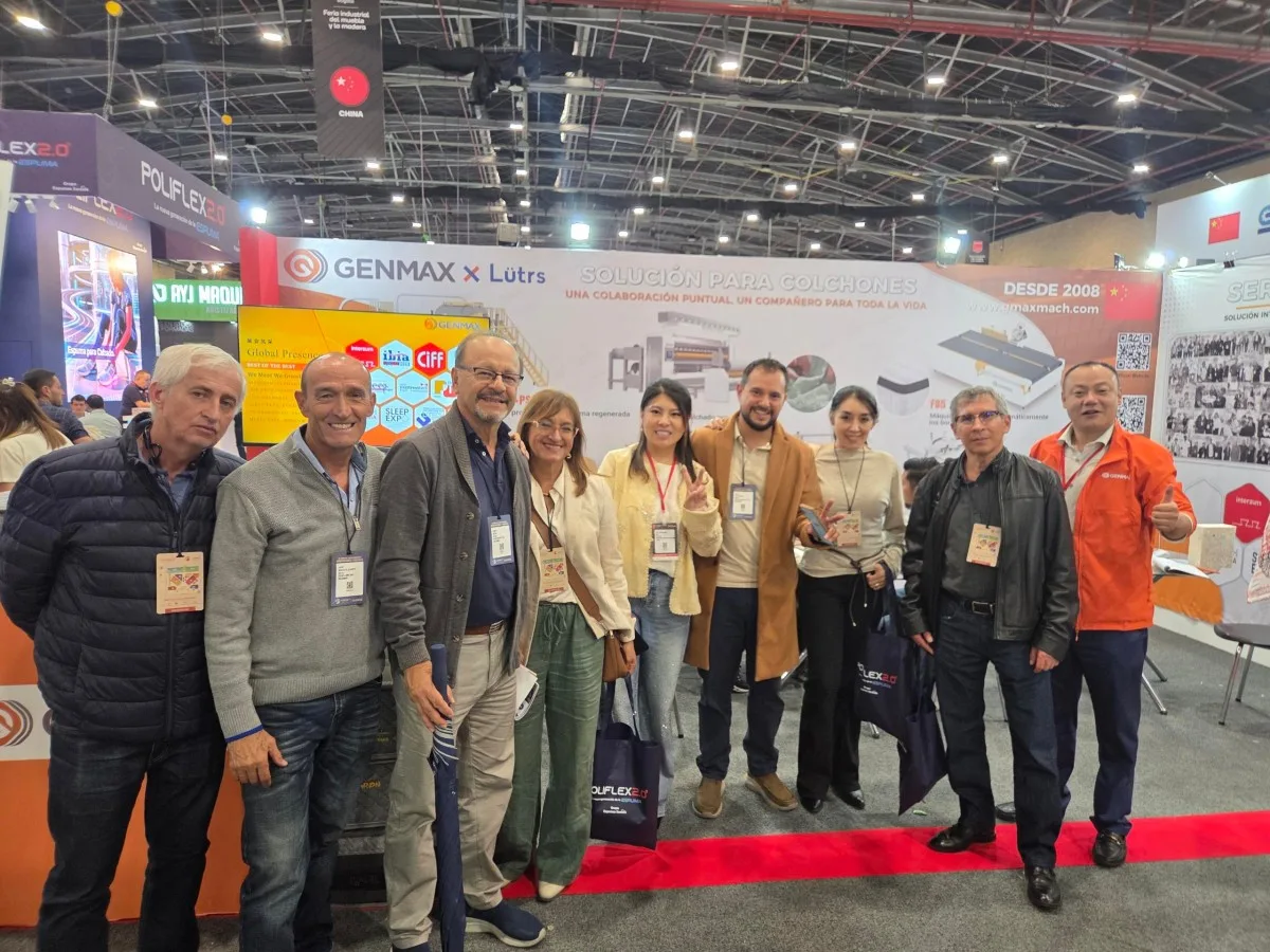 GENMAX sales team and industry visitors posing together in front of the GENMAX x Lütrs "Solución Para Colchones" booth backdrop at Interzum Bogotá 2026, Corferias, Colombia
