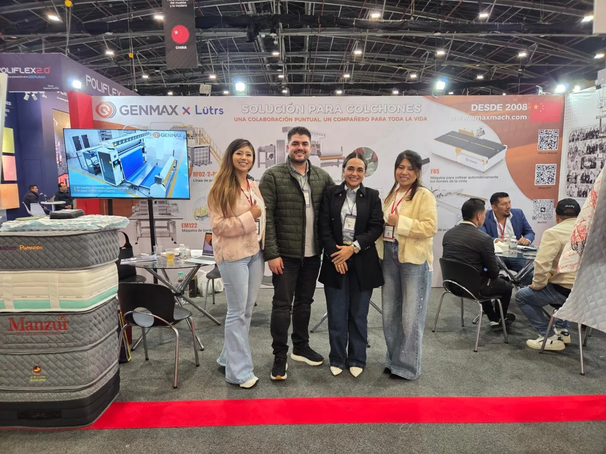 Four people — one male visitor and three GENMAX team members — posing together in front of the GENMAX x Lütrs "Solución Para Colchones" booth, with mattress displays and product demonstration screen visible, Interzum Bogotá 2026
