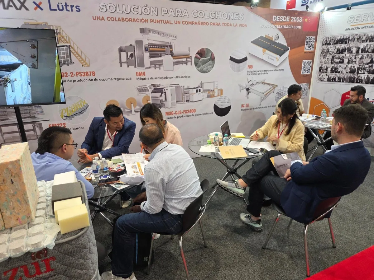 GENMAX team members posing with a visitor in front of the GENMAX x Lütrs booth display, with mattress samples and product demonstration screen visible, Interzum Bogotá 2026