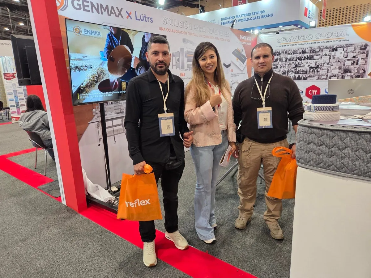 Two industry visitors holding orange Reflex bags posing with a GENMAX team member in front of the GENMAX x Lütrs "Solución Para Colchones" booth backdrop at Interzum Bogotá 2026