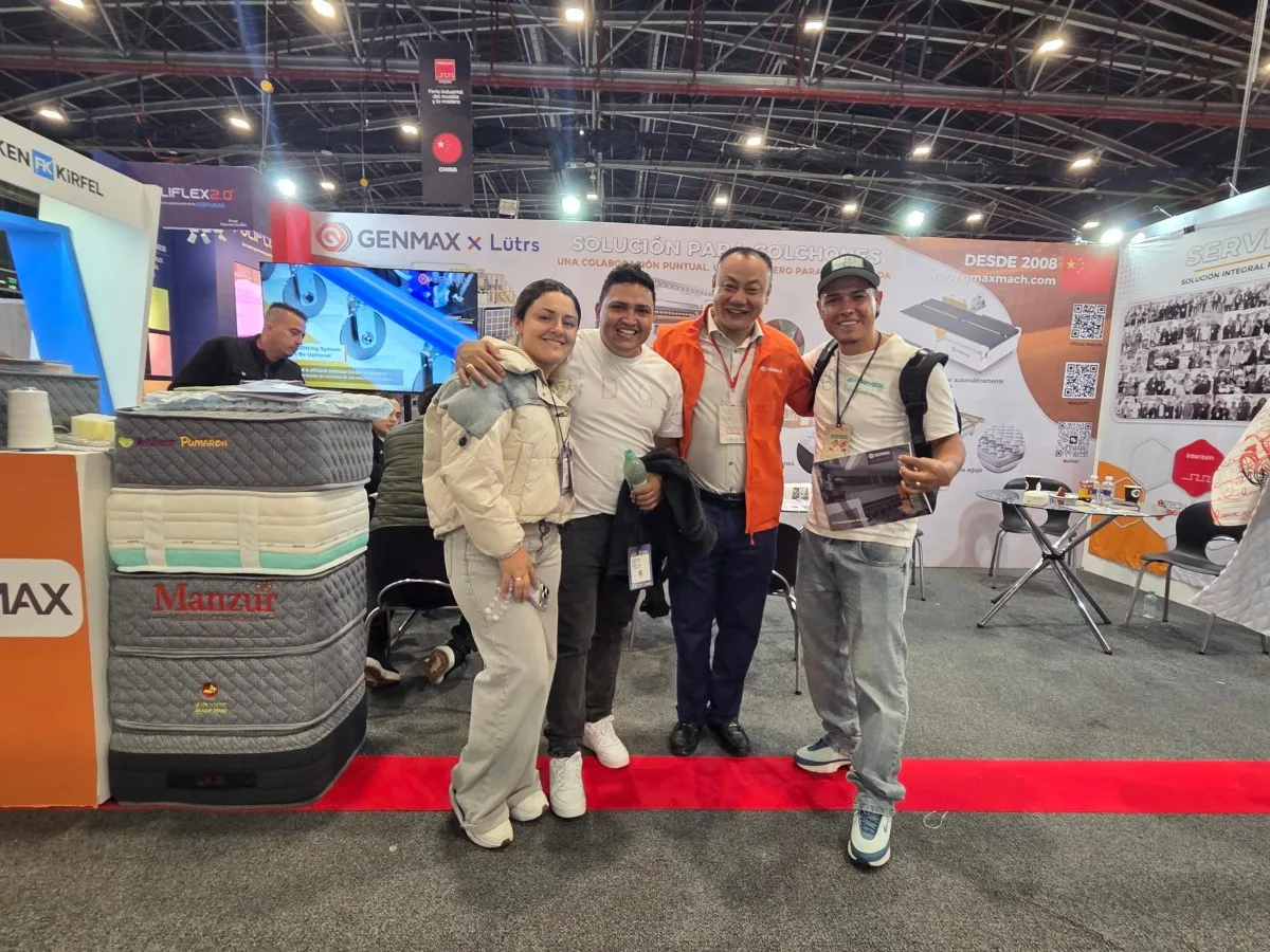GENMAX sales representative in orange jacket posing warmly with three young industry visitors in front of the GENMAX x Lütrs booth, with Manzur branded mattress displays visible, Interzum Bogotá 2026